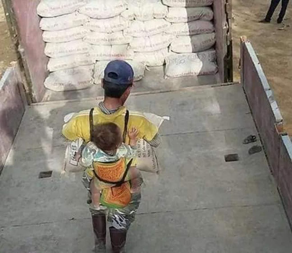En su espalda carga al bebé en una especie de morral mientras levanta bolsas con sus brazos.