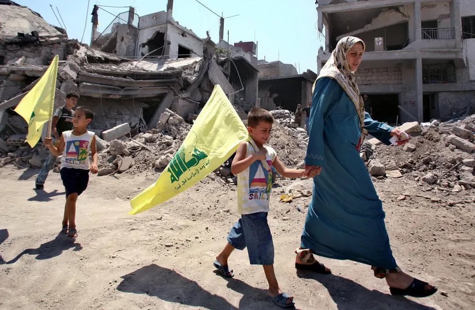 Dos niños con su madre caminando por las calles destruidas de Líbano cargando banderas de Hezbolá.