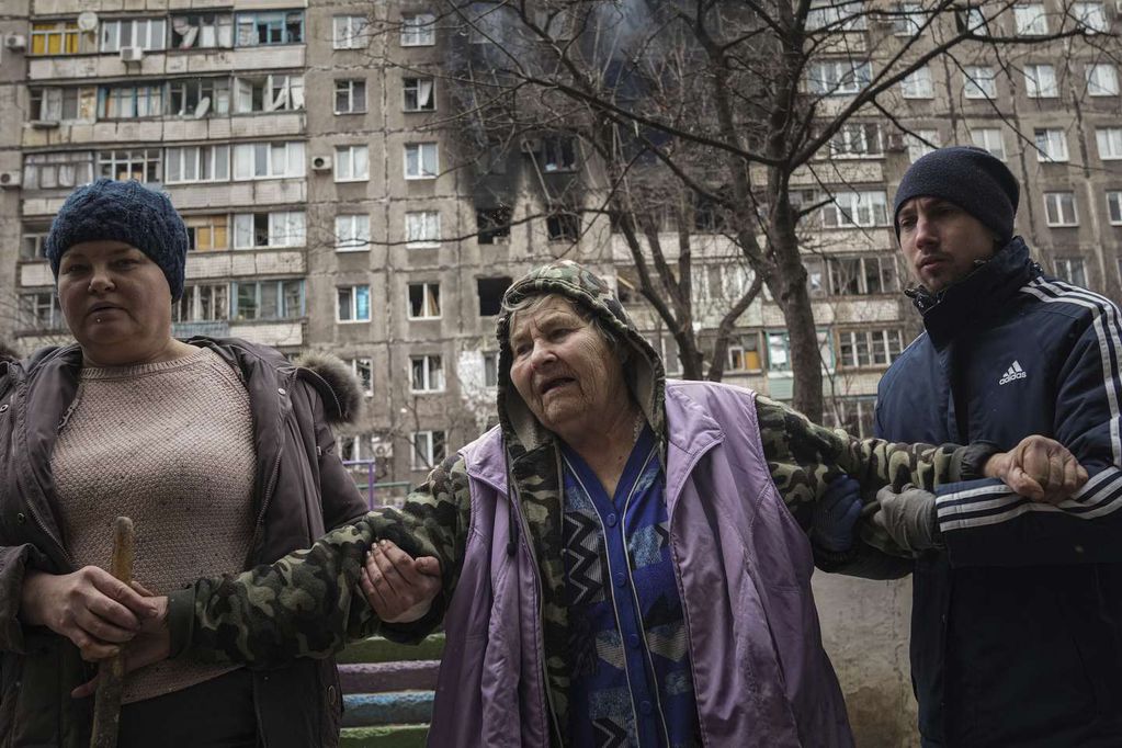 Gente ayudando a una anciana a caminar por la calle junto a un edificio de apartamentos alcanzado por proyectiles en Mariúpol, Ucrania, el lunes 7 de marzo de 2022. / Foto: AP