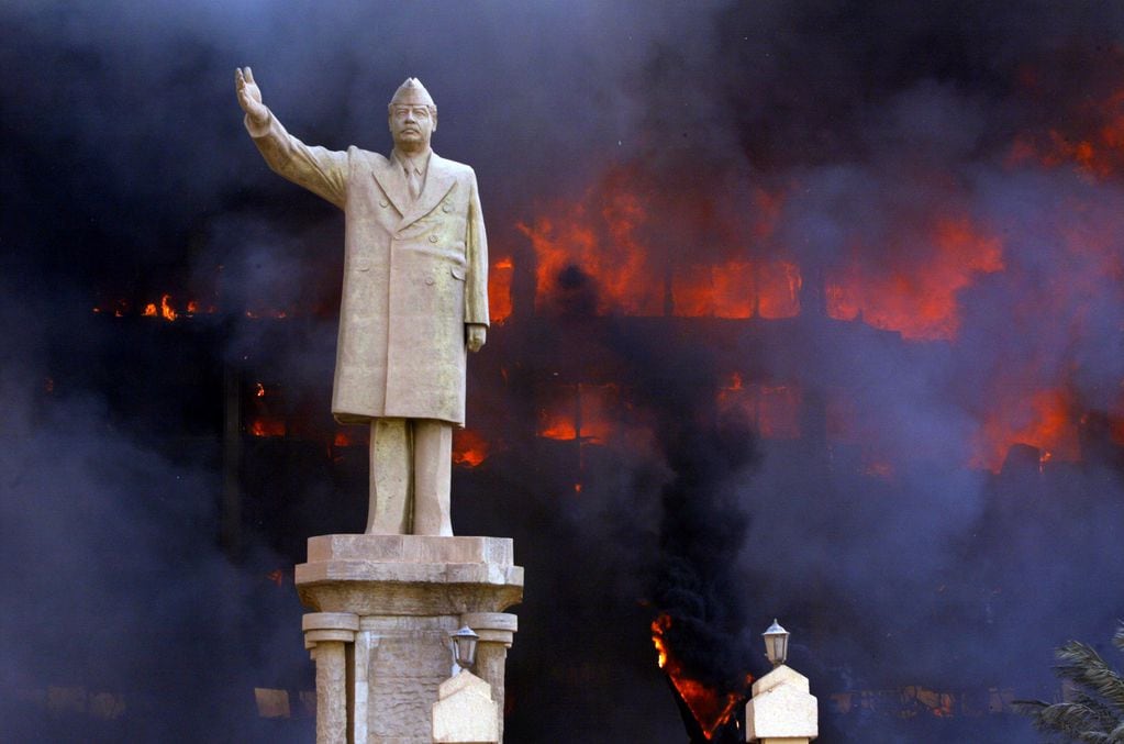 Una estatua de Sadam Husein antes de ser destruida mientras arde en llamas un edificio público.