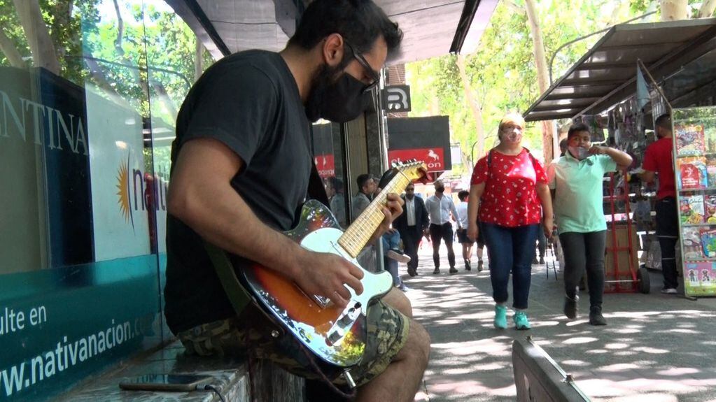 El músico Diego Lorca, sentado afuera de una de las sedes del Banco Nación, toca su guitarra eléctrica mientras los transeúntes pasean por la calle.
